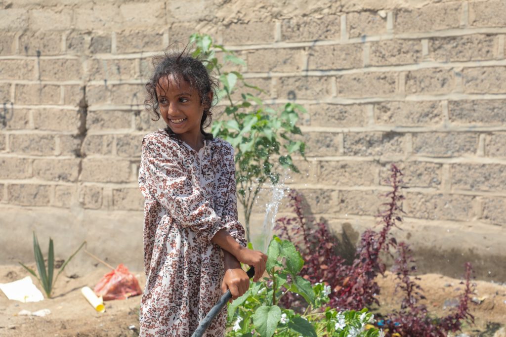 Smiling girl watering plants with a hose in Al-Khukhah, Yemen, thanks to improved water access