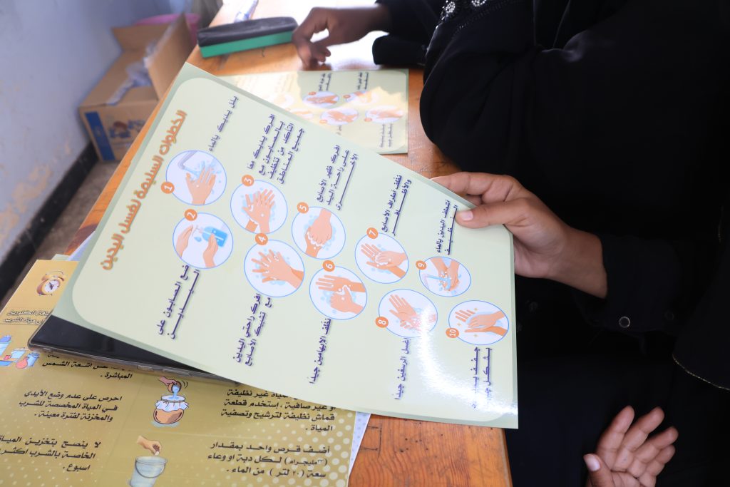 Woman holding an Arabic-language poster illustrating ten handwashing steps during a hygiene awareness session in Yemen