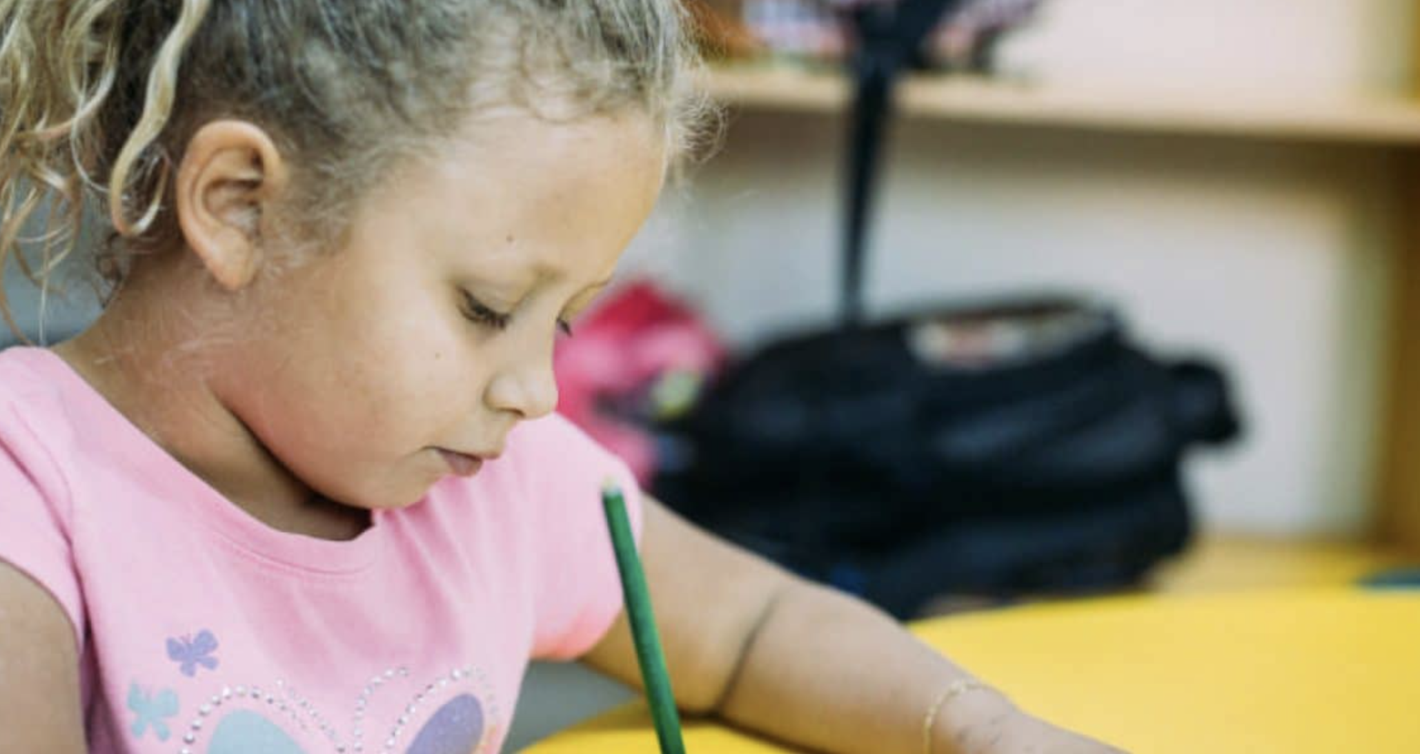 A young girl with curly hair concentrating on drawing or writing with a pencil at a table