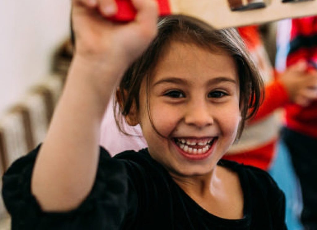 A young girl laughing and holding up a wooden toy with other children playing in the background