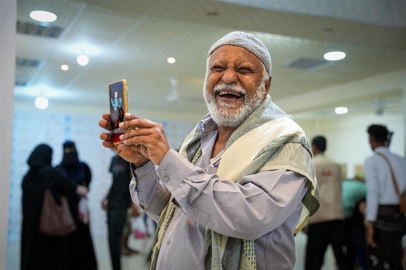 Älterer jemenitischer Mann mit traditioneller Kappe lacht fröhlich und hält ein Smartphone hoch bei einer Veranstaltung