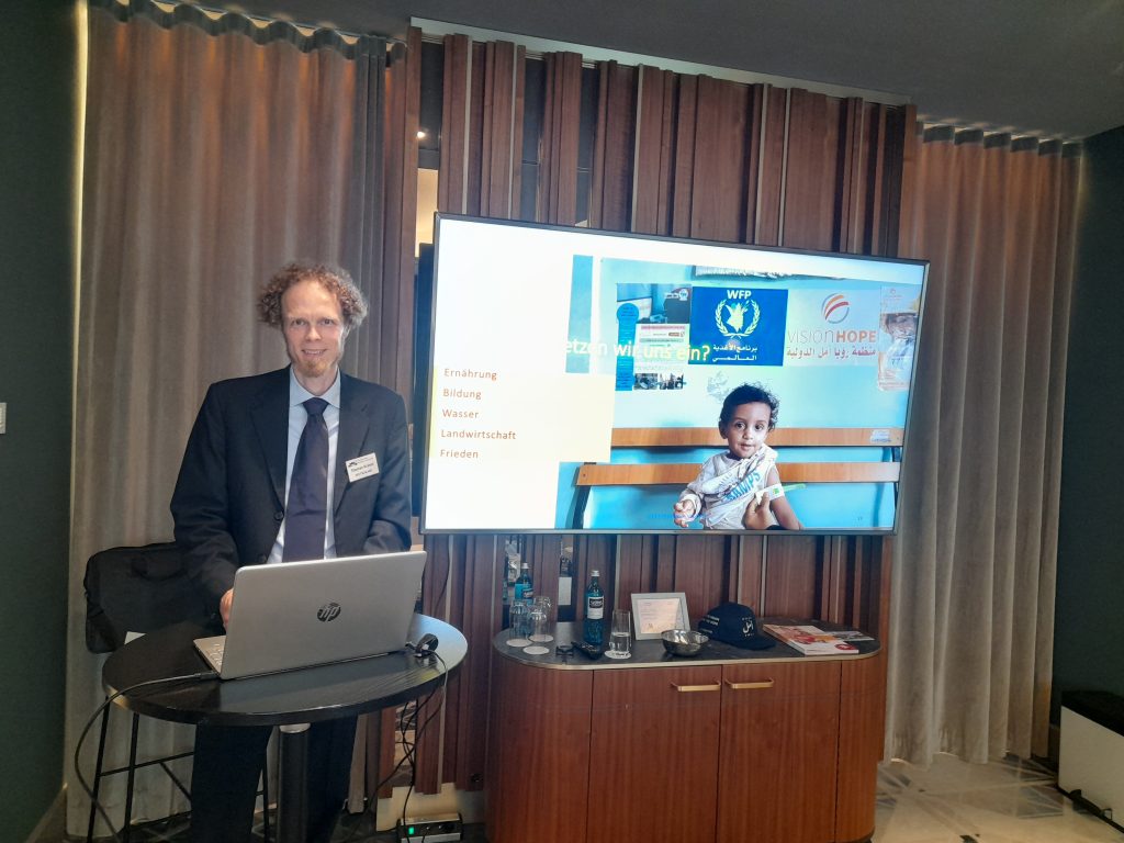 Speaker presenting Vision Hope's work to German humanitarian organizations at a Berlin networking event, with a screen showing project themes and a photo of a child from Yemen