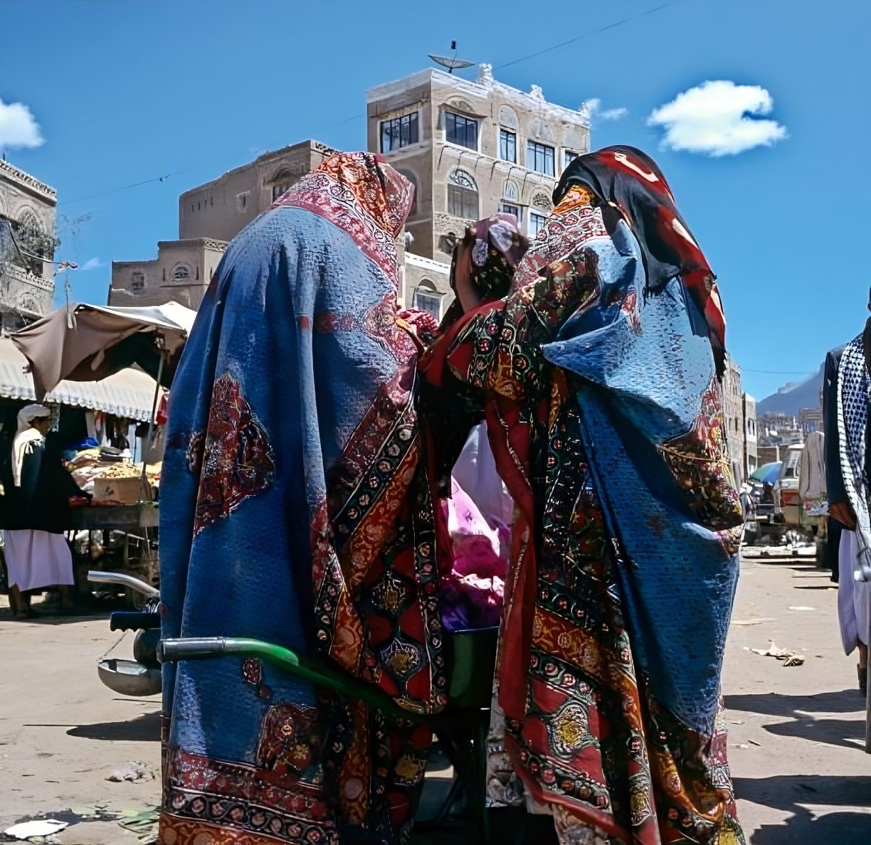 Two Yemeni women wearing colorful traditional sitara wraps at a Sana'a market, with old-town tower houses behind